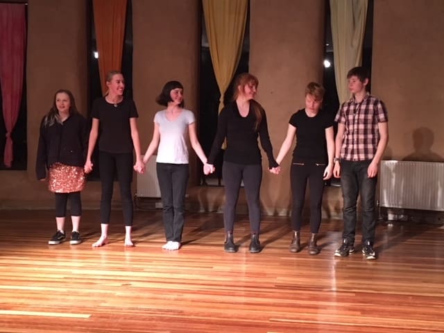 Students stand together on stage, holding hands to celebrate the courage and connection of sharing their original voices.