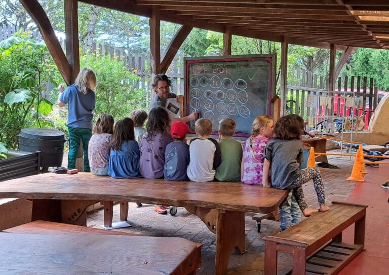 Students gather on a timber table, honouring natures rhythmic lessons within a bush-centred outdoor classroom.