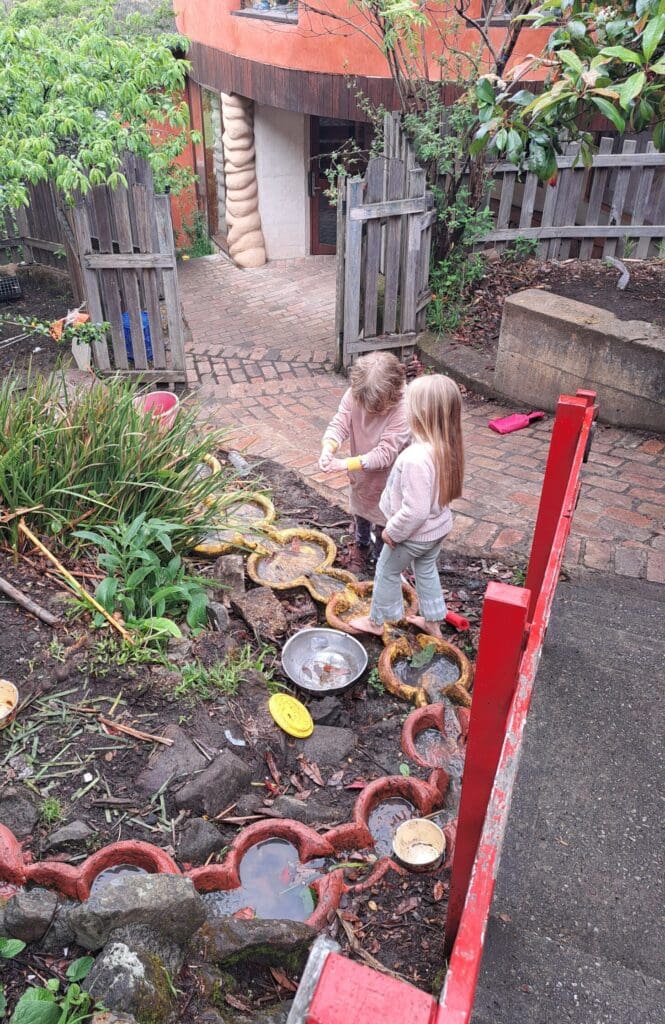 Barefoot children discover the rhythm of water and earth, cultivating wonder within Kindlehills lush garden environment.