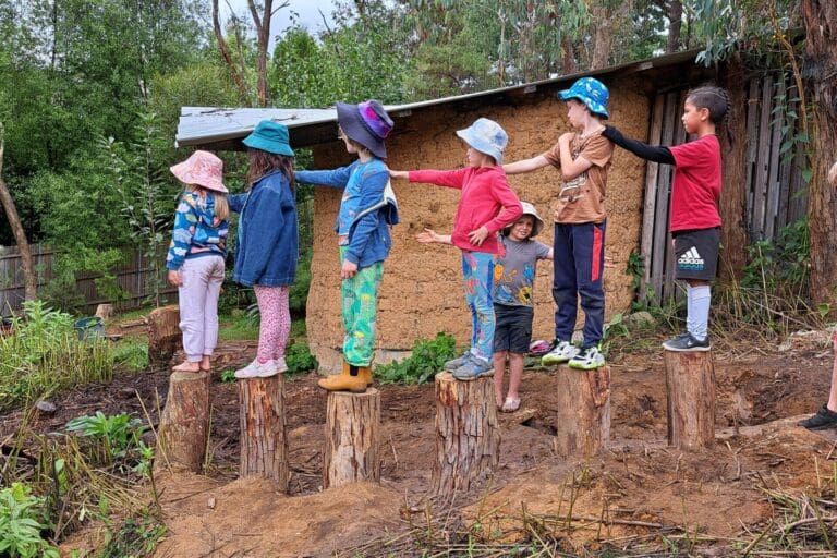 Kindlehill children balance together on stumps, supporting one another and connecting to Country within their bushland environment.