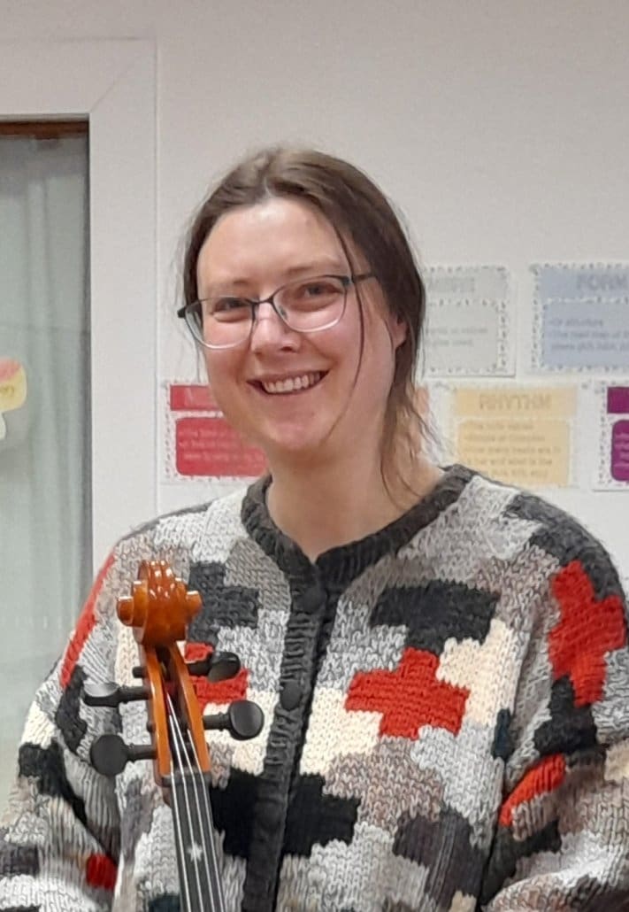 A Kindlehill teacher smiles warmly holding a cello, embodying artistic expression and the rhythmic community breath of music.