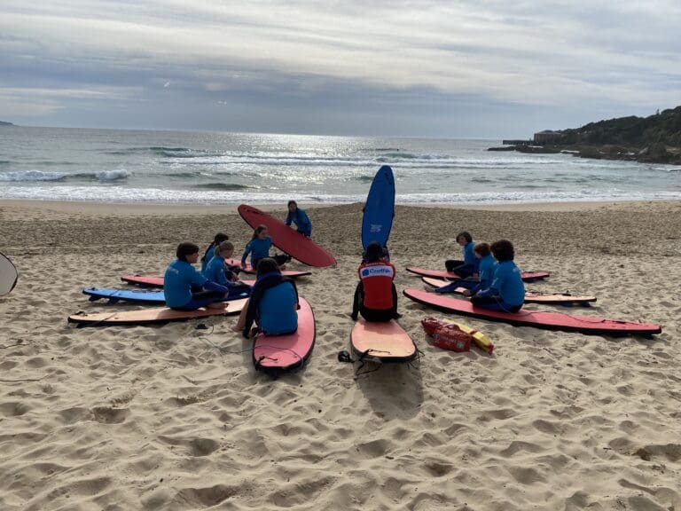 Students gather in a circle on surfboards, cultivating resilience and community spirit through immersive, nature-based learning by the ocean.